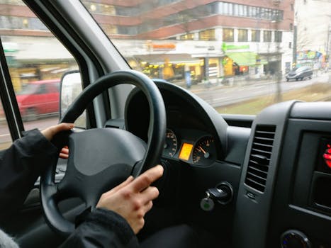 Hands on a steering wheel driving a van through an urban area with shops visible outside.